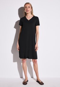 Schwarzes kurzärmliges Kleid mit V-Ausschnitt, aus glattem Stoff, mit lockerer Passform und knielangem Saum. Getragen mit schwarzen Mesh-Schuhen.