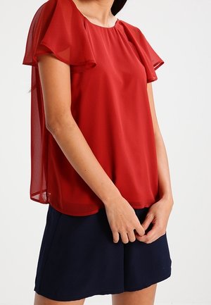 Kobieta w czerwonej bluzce z krótkim rękawem, z przezroczystą tkaniną na rękawach i plecach, w parze z granatowymi szortami, stojąca z rękami złożonymi razem.