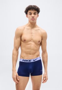 Άνδρας χωρίς πουκάμισο με σγουρά μαλλιά, ντυμένος με μπλε-σκούρο boxer της Nike με γαλάζια μέση, στέκεται μπροστά σε απλό λευκό φόντο.