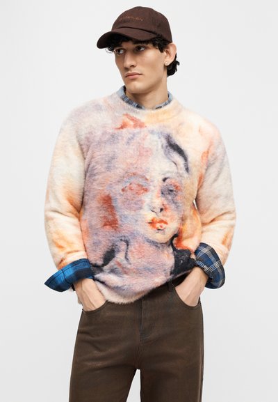 Πλεκτή, πολύχρωμη ζακέτα με αφηρημένο πορτρέτο σε αποχρώσεις πορτοκαλί, ροζ και μπλε, συνδυασμένη με καρό πουκάμισο.