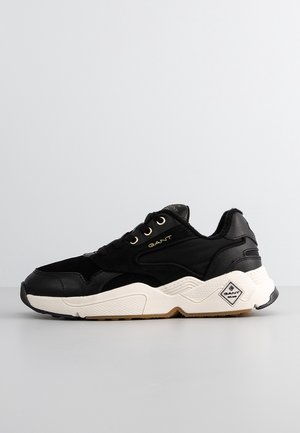 Czarny sneaker GANT z białą teksturowaną podeszwą, złotymi oczkami i logo po bokach oraz na pięcie, prezentowany na jednolitym szarym tle.
