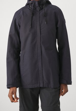 Person iført en sort hættetrøje med snore, lange ærmer og forlommer, kombineret med sorte shorts.