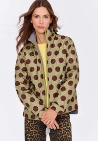 Zelená prešívaná bunda s veľkými hnedými bodkami, sivou podšívkou, vysokým golierom a zipsom. Nosí sa cez žltý top so zvieracím vzorom a nohavicami.
