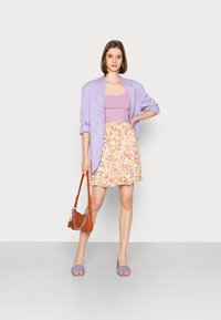 Blazer oversize de lavanda, top canelado lilás, saia de padrão animal em laranja e amarelo, sandálias tecidas roxas, bolsa de ombro castanha com corrente.
