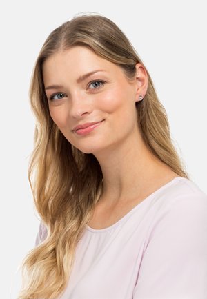 Runde Steckerohrringe mit klaren und bunten Edelsteinen, die in einem silberfarbenen Metall gefasst sind. Das Modell hat lange, gewellte blonde Haare.