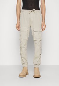 Béžové cargo kalhoty s elastickým pasem a lemy, s několika bočními kapsami. Hladká tkanina s volným střihem. Dokončeno hnědými botami.
