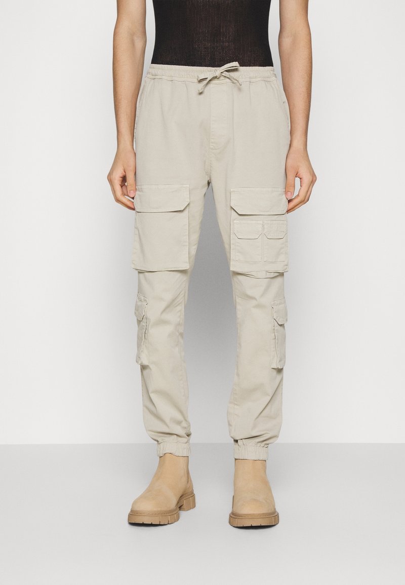 Béžové cargo kalhoty s elastickým pasem a lemy, s několika bočními kapsami. Hladká tkanina s volným střihem. Dokončeno hnědými botami.