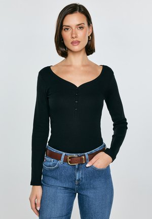 Rövid barna hajú nő, fekete hosszú ujjú, gombos felsőt, kék farmert és barna övet visel, egyik kezét a zsebében tartva áll.