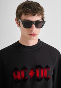 Zwarte sweatshirt met rood AC/DC-logo, gecombineerd met hoekige zwarte zonnebril. Model heeft krullend haar en een neutrale uitdrukking.