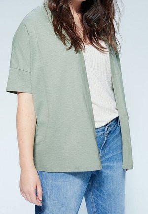 Femme portant un cardigan court à manches courtes vert clair, ouvert sur un haut blanc côtelé et un jean bleu, sur un fond uni.