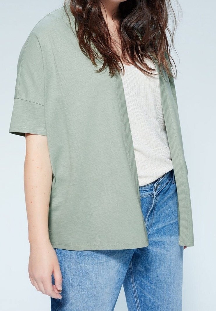 Femme portant un cardigan court à manches courtes vert clair, ouvert sur un haut blanc côtelé et un jean bleu, sur un fond uni.