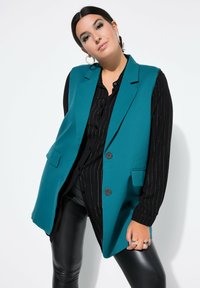 Blazer verde azulado con un corte entallado, solapas con muesca, dos bolsillos frontales y botones negros, llevado sobre una blusa negra a rayas verticales y pantalones de cuero.
