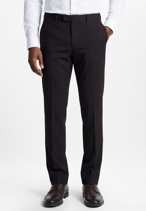 Mann trägt schwarze formelle Hose, weißes Hemd und dunkelbraune Lederschuhe, steht mit einer Hand in der Tasche.