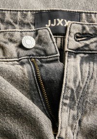 Juoda ir balta džinsinė kelnė su užtrauktuku, sidabriniu mygtuku ir audinio etikete, užrašyta "JJXX". Medžiagos tekstūra atrodo šiek tiek nusidėvėjusi.