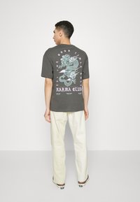 Gray cotton t-shirt with short sleeves, featuring a large white and light blue dragon graphic and text on the back, paired with cream-colored pants.