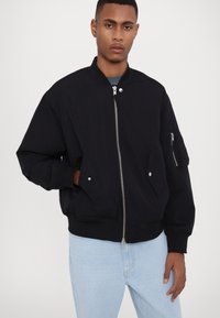 Black bomber jacket made of lightweight fabric, featuring a front zipper, side pockets, and ribbed cuffs. Worn over light blue jeans.