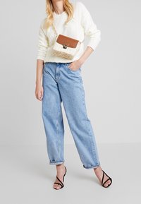 Femme portant un pull blanc en maille torsadée, un jean taille haute bleu clair, des sandales noires à lanières et un petit sac bandoulière tissé marron et blanc.