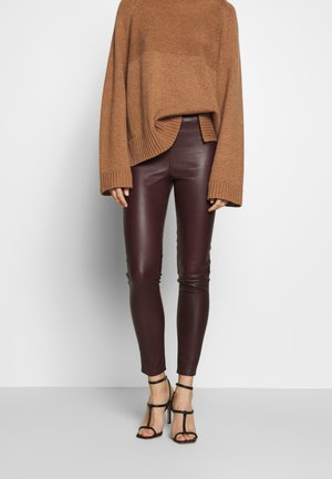 Vrouw draagt een bruine oversized gebreide trui, donkerbruine leren nauwsluitende broek en zwarte sandalen met hoge hakken en enkelbandjes.