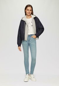 Granatowa kurtka bombowiec z szarym kapturem, jasnoniebieskie obcisłe dżinsy i białe sneakersy. Posiada swobodny krój z zamkiem z przodu oraz graficznym nadrukiem.