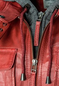 Rote Lederjacke mit grauem Kapuzen, metallenen Reißverschlüssen, geflochtenen Kordeln und einer Brusttasche. Strukturierte Oberfläche mit sichtbaren Nähdetails.