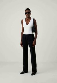 Sieviete ģērbta balta bezpiepeša V-veida augšdaļā, melnās platas bikses, melnas norādītas kurpes, saulesbrilles un nesot melnu pleca somu.