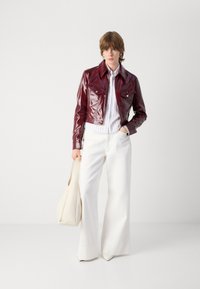 Burgundy patent leather cropped jacket with buttoned pockets, paired with white pleated blouse and wide-leg white trousers. Beige handbag.