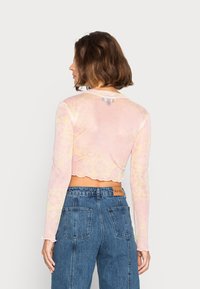 Top curto de mangas compridas em tecido rosa transparente com padrão floral. Apresenta um decote redondo e uma bainha recortada. Combinado com jeans de cintura alta.