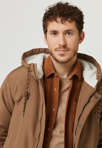 Giacca marrone con foderatura in sherpa bianco, dotata di chiusura con zip e cappuccio regolabile con cordino. Indossata sopra una camicia in velluto a coste color ruggine.