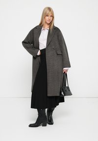 Grijze wollen jas met een enkele knoop, oversized ontwerp, gecombineerd met een lichtpaarse blouse en een zwarte geplooide rok. Inclusief zwarte handtas en laarzen.