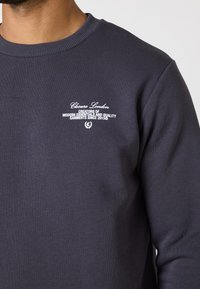 Mørkeblå sweatshirt lavet af bomuld. Har en ribbet halsudskæring og en broderet logo i hvid på brystet. Glat tekstur. Minimalistisk design.