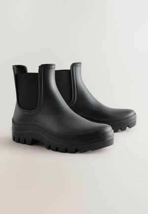 Bottes de cheville en caoutchouc noir avec des panneaux latéraux texturés et une semelle épaisse et robuste, présentant des orteils arrondis et une légère élévation au niveau du talon.