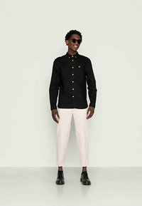 Άντρας φορώντας μαύρο πουκάμισο με κουμπιά, μπεζ κοντές παντελόνες, μαύρα παπούτσια και γυαλιά ηλίου, αφού στέκεται μπροστά από απλό ανοιχτό φόντο.