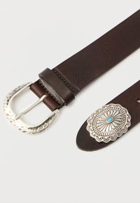 Pruun nahast vöö, millel on hõbedane haamriga viimistletud pandl ja dekoratiivne hõbedane ots, millel on reljeefne lillemuster ja türkiissinine aksent.
