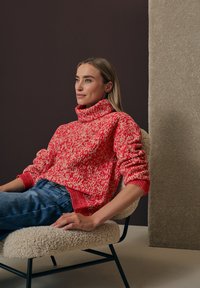Un suéter de punto grueso en tonos rojos y crema con un cuello alto, combinado con jeans de mezclilla azules. El entorno presenta suaves texturas en beige.