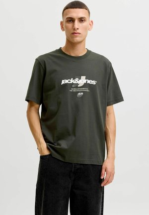 Ung mand med kort hår iført en mørkegrøn Jack & Jones T-shirt med grafik og sorte bukser, stående med den ene hånd i lommen.