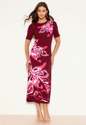 Frau in einem taillierten, burgunderroten Midikleid mit großen pinken Blumenmustern, kurzen Ärmeln und beigen, riemchenartigen High-Heel-Sandalen vor neutralem Hintergrund.