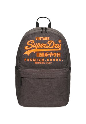 Szary plecak z materiału z pomarańczowym haftowanym logo, wyposażony w przednią kieszeń na zamek błyskawiczny oraz regulowane czarne szelki. Gładka tekstura.