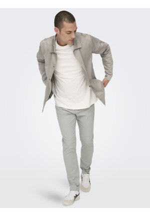 Veste gris clair sur une chemise blanche, associée à un pantalon gris clair et des baskets blanches. La veste a un design simple et des détails de boutons.
