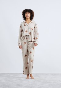 Lichtbeige pyjamaverset met een knoopsluiting bovenkant en broek met trekkoord, met een herhaald bruin teddybeerpatroon. Zachte stoftextuur.