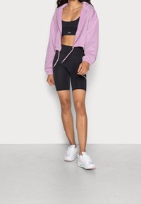 Růžová krátká mikina s kapucí, černá sportovní podprsenka, černé cyklistické šortky a bílé tenisky s pastelovými akcenty; hladká tkanina a přiléhavý střih.