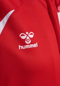 Tissu de sport rouge avec logo blanc Hummel représentant une abeille stylisée et un détail de chevron blanc partiel à côté d'une fermeture éclair rouge.