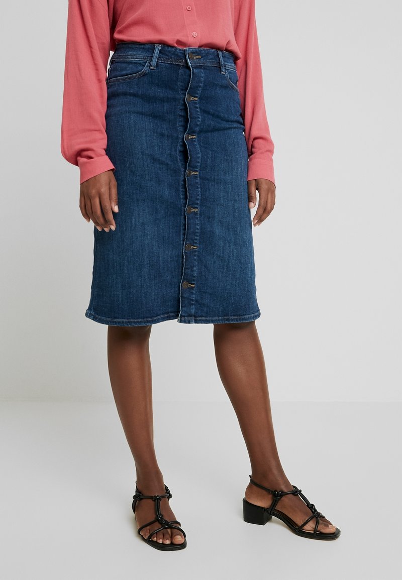 Jupe en jean bleu de longueur moyenne avec boutons sur le devant, portée avec une chemise rose à manches longues et des sandales noires à talons avec lanières.