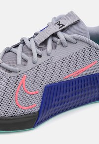 Scarpa sportiva grigia con tomaia in mesh, dettagli rosa e suola texturizzata blu. Presenta un sistema di allacciatura tradizionale e un collo imbottito per il comfort.