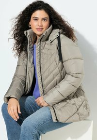 Giacca puffo beige trapuntata con cappuccio, dettagli neri, chiusura frontale con zip e polsini elastici, indossata sopra una maglietta viola e jeans blu.