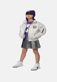 Veste bomber argentée, bonnet violet et t-shirt, jupe métallique, baskets montantes blanches avec accents noirs et logos.