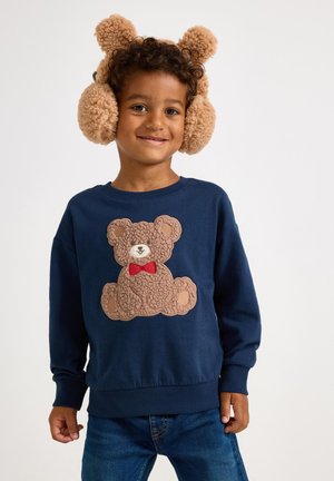 Marinblå sweatshirt med ett texturerat brunt nallebjörnsmotiv och röd fluga. Bruna fluffiga öronvärmare med runda former. Barnmodell som ler.