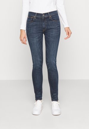 Persoon die een donkerblauwe skinny jeans en witte sneakers draagt, staand tegen een effen lichte achtergrond.