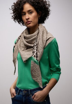 Femme aux cheveux bouclés portant un pull vert, un jean bleu et une écharpe beige à motifs drapée autour du cou, les mains dans les poches, fond neutre.