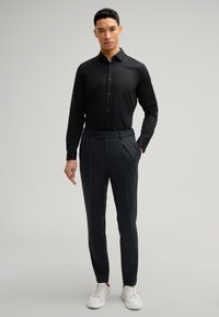 Zwarte langemouwen shirt met knopen, gecombineerd met donkergrijze broek met plooien en een riem, afgewerkt met witte sneakers.