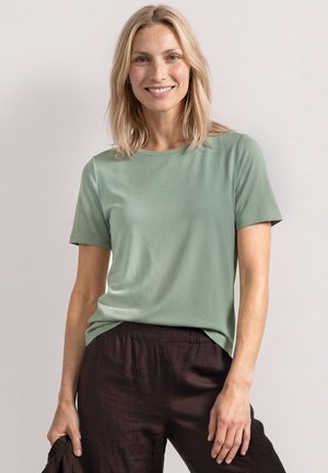 Kobieta o blond włosach do ramion, ubrana w jasnozieloną koszulkę z krótkim rękawem i ciemnobrązowe spodnie, uśmiecha się na jednolitym tle.
